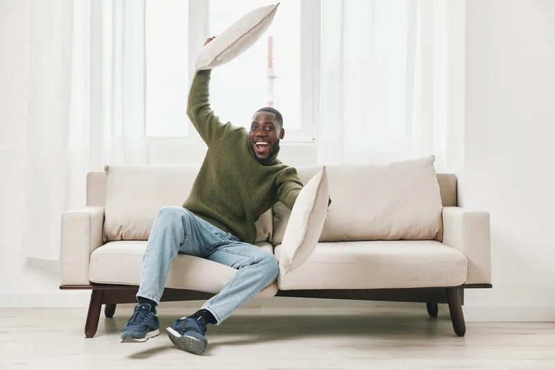 A joyful Black man playfully raising cushions on a modern beige sofa, expressing happiness and excitement in a bright room, perfect for lifestyle and home decor themes