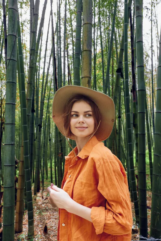 Woman bamboo forest hat portrait smile, wearing a wide-brim hat and orange shirt in a serene three-quarter portrait on a leaf-strewn path among tall green stalks in daylight.