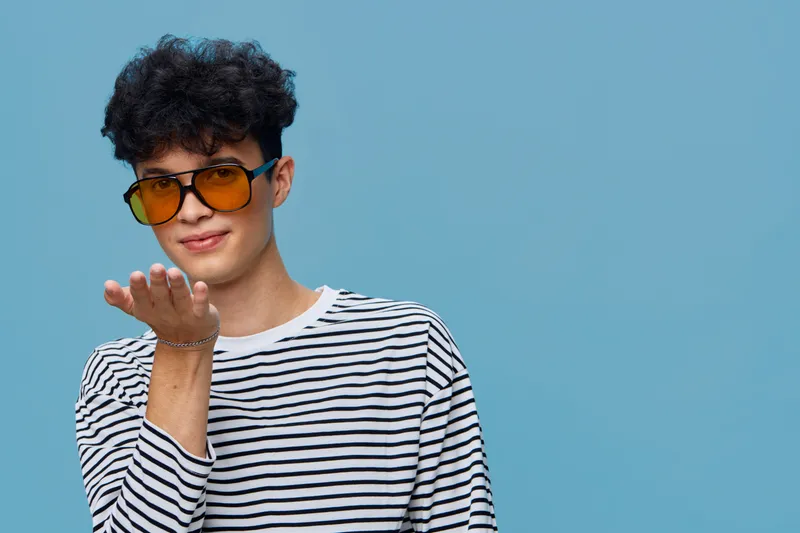 young man wearing yellow sunglasses and striped shirt blowing a kiss with blue background, casual style and playful mood in studio lighting