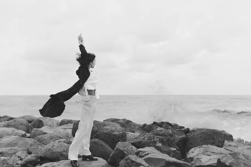 Silhouette of a woman on a rocky shoreline, wearing light pants and a flowing jacket, gazing at the sea as a sweeping wind lifts fabric, a moody monochrome scene of movement and solitude