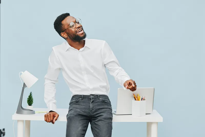 Confident smiling young man with glasses, casual white shirt and jeans standing at modern white office desk isolated on light blue background, relaxed pose, promoting positive professional atmosphere.