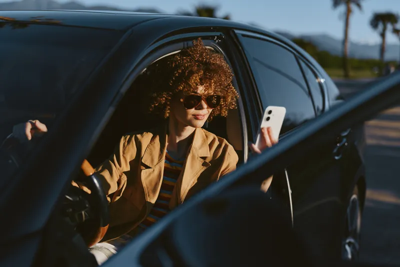 Young woman with curly hair sits in a car looking at her phone, wearing a tan blazer and sunglasses, captured in warm retro film tones during a sunlit city drive with a relaxed, contemplative mood