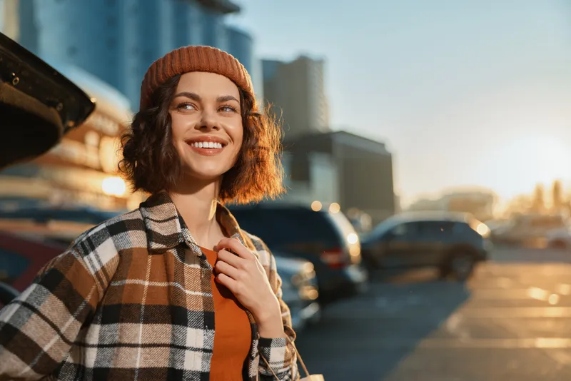Woman smile urban golden candid lifestyle beanie parking. Young woman smiling in a plaid coat at golden hour in a city parking, authenticity, mindful living, emotional storytelling, relaxed portrait.