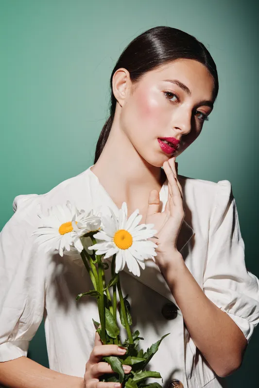 Young woman with glossy skin and red lips poses with daisies on a green background wearing a white blouse. Concept of beauty glamour fashion creative modern style portrait.