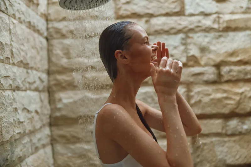 young woman enjoying outdoor shower with stone wall background, wet hair, and relaxed joyful expression in natural light setting.