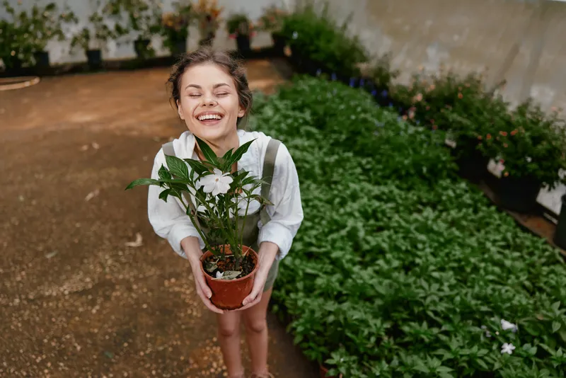 Happy girl holding a potted plant in a greenhouse, showcasing joy and connection with nature in a vibrant, green environment filled with flowers