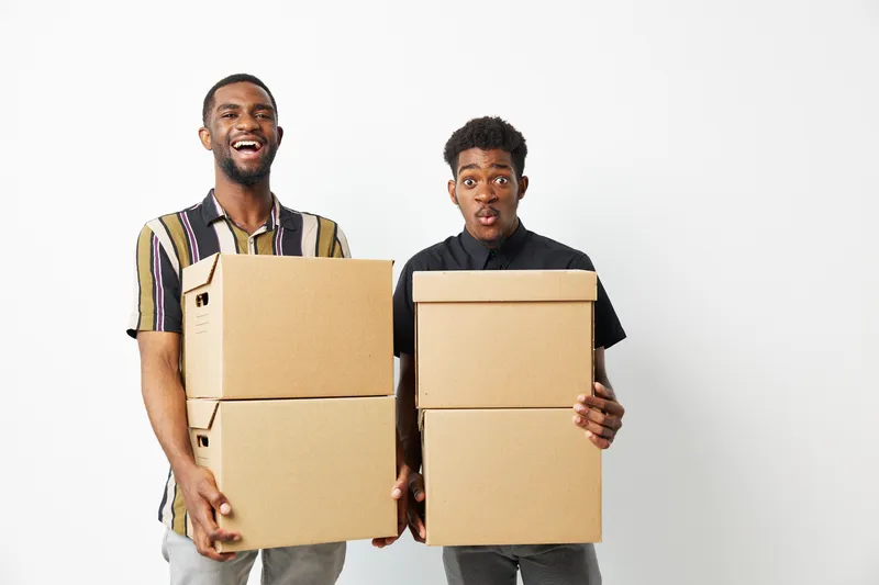 Two smiling black men joyfully holding cardboard boxes, showcasing friendship and collaboration in a bright setting with a minimalist background.