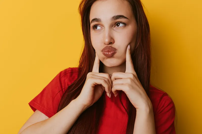 Young woman with long hair wearing red shirt posing with fingers on cheeks and thoughtful expression against yellow background. Beauty, emotion, portrait and casual style with natural look.