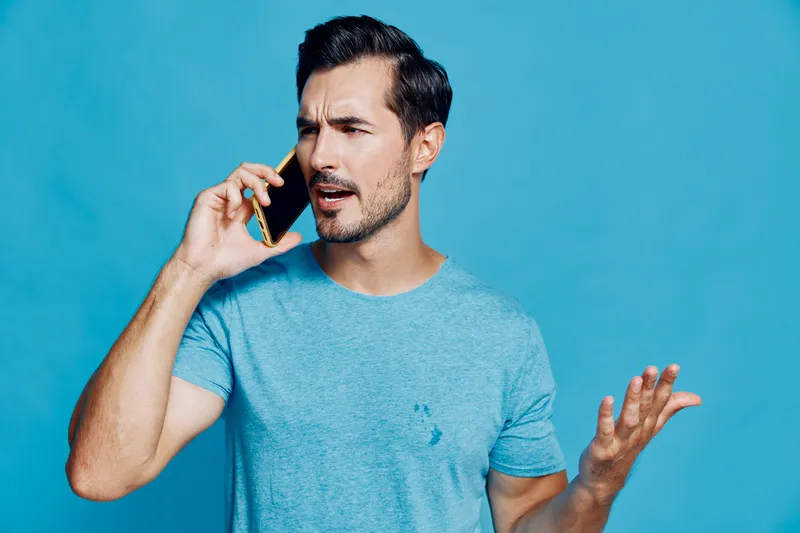 Confused man talking on mobile phone with puzzled expression, gesturing with hand, isolated on bright blue background in studio portrait. Communication concept.