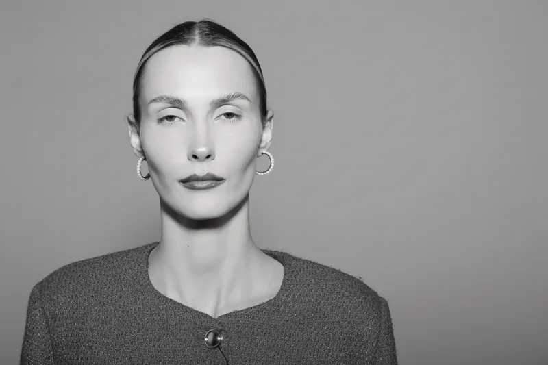 Tall slender Caucasian woman with pale skin and slicked back hair wears textured jacket and hoop earrings, posing with neutral expression on gray background.