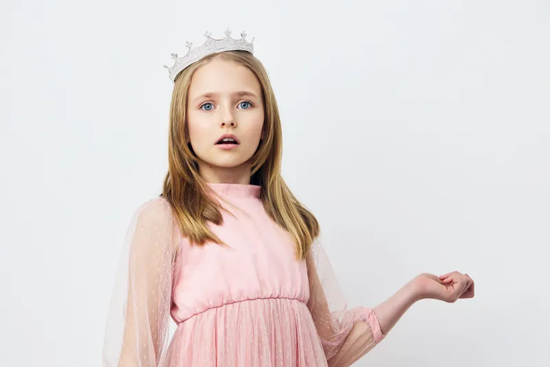 Young girl in a pink dress and crown, expressing surprise and wonder against a neutral background, symbolizing childhood dreams and imagination
