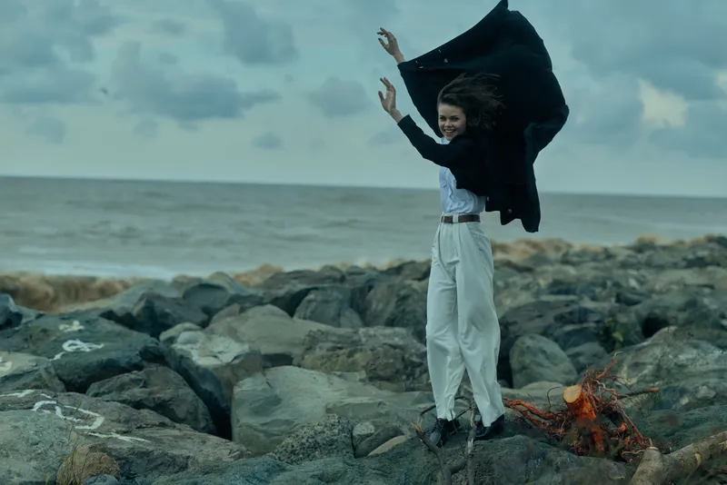 Woman wearing a flowing cape on a rocky shore, wind lifting fabric, dramatic outdoor scene by the sea with dynamic movement and bold fashion.
