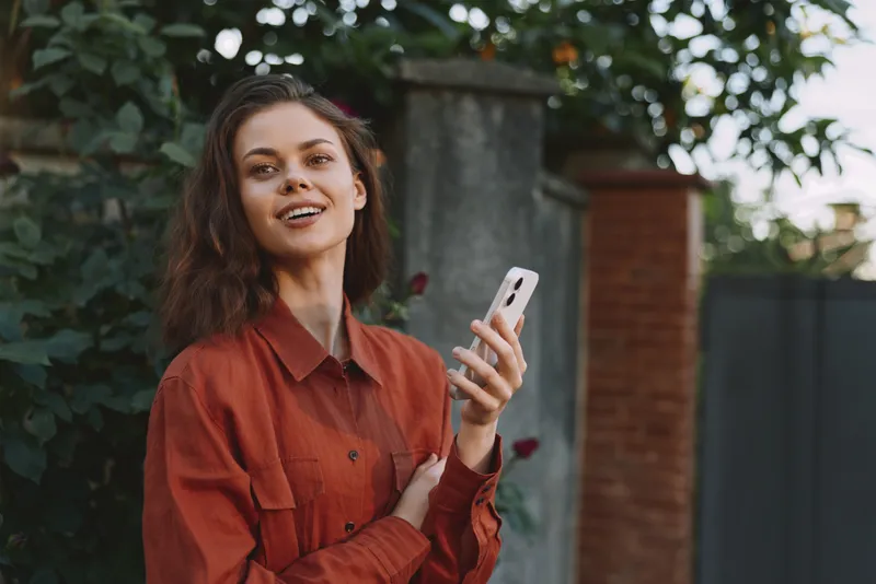 Woman outdoors, smiling, holding a smartphone in casual fashion, natural light, candid lifestyle moment with greenery in the background, relaxed pose, vibrant colors, sunny breeze