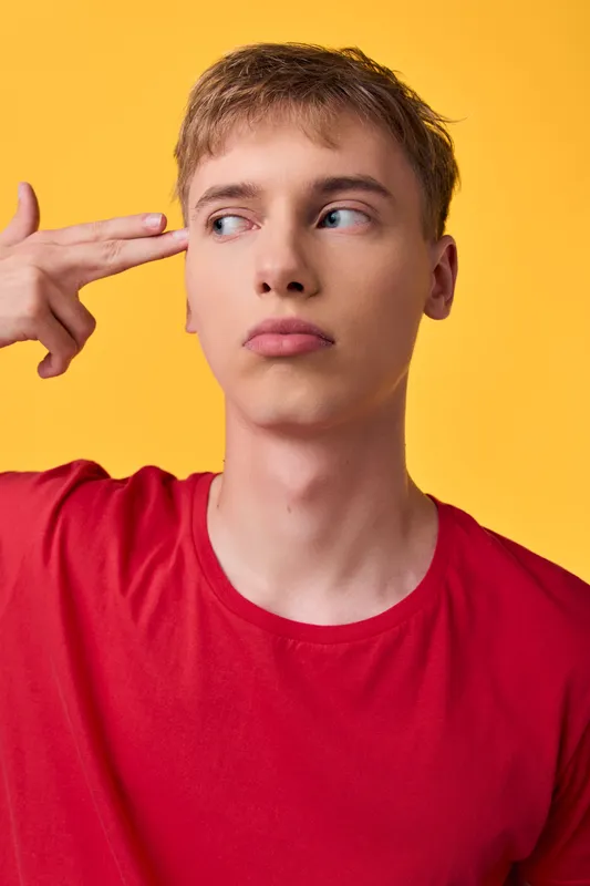 Thinking man wearing a red shirt against a bright yellow backdrop in studio, showing a curious expression and a finger to temple, conveying focus, creativity and mood