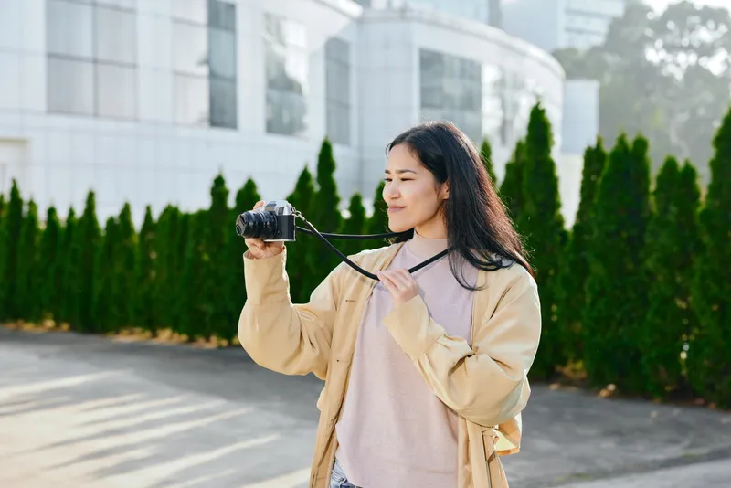 Outdoor camera scene shows a woman holding a compact camera, wearing a beige jacket and pink sweater, standing beside modern glass buildings and trimmed hedges