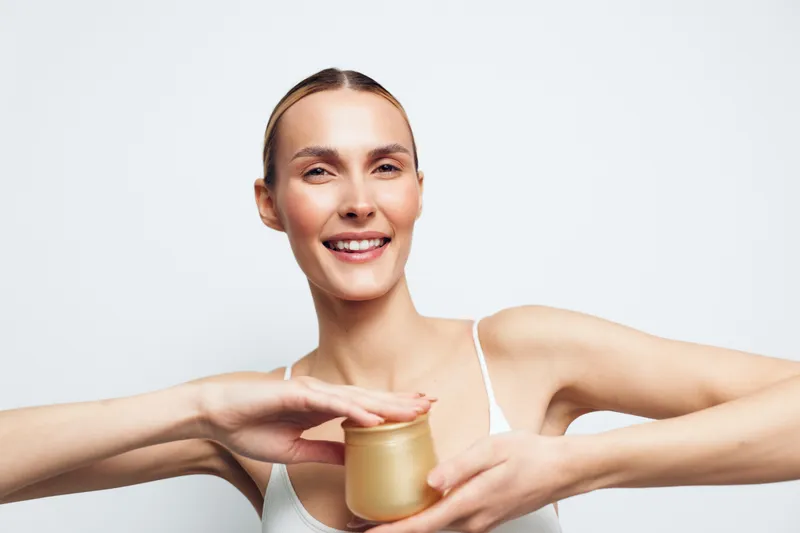 Young woman with blonde hair wearing a white tank top holds a jar of cream and applies it to her face, smiling warmly in a bright studio setting.