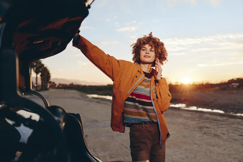 Woman outdoors by a car at sunset, smiling in warm evening light, curly hair loose, wearing a jacket, casual travel lifestyle, open road background, calm mood, golden sky, distant fields