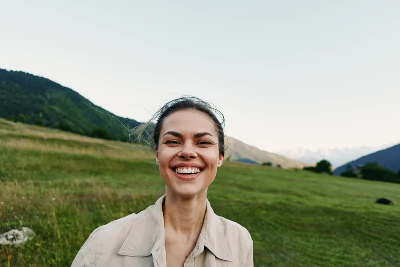 Happy woman outdoors smiling broadly with natural background of green hills and clear sky, casual outfit, fresh air, relaxed mood, lifestyle concept