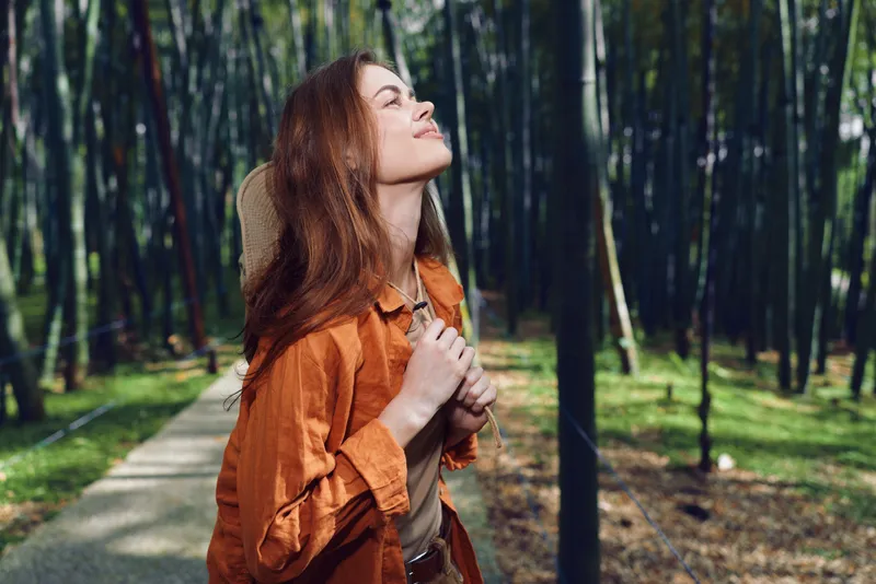 Woman with backpack on forest path hiking and enjoying nature with eyes closed and serene smile, sunlight filtering through trees for a peaceful outdoor travel moment.