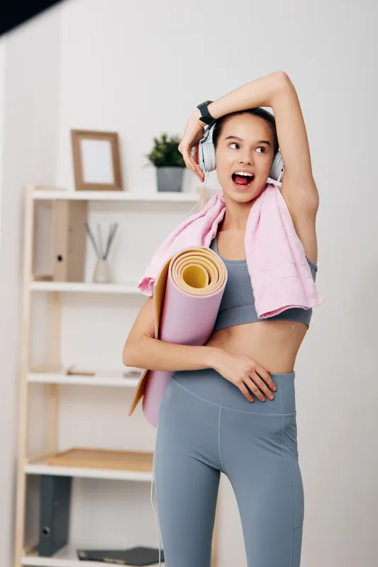 Female fitness enthusiast wearing workout apparel, headphones, and a towel after exercise, holding a yoga mat in a bright home gym, ready for training.