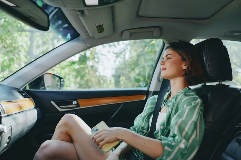 Woman sits in a car interior, wearing a green striped shirt, buckled up, reading a notebook as sunlight spills through the window.