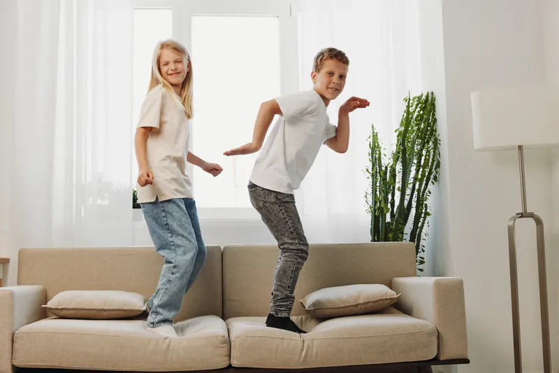 Two happy children jumping on a beige couch in a bright living room. Playful brother and sister enjoying active fun at home with natural light and casual clothing.