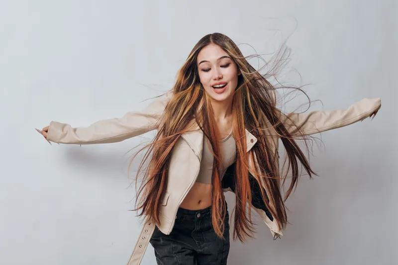 Young woman with long hair dancing and smiling wearing casual jacket and top against light background for lifestyle and fashion portrait.