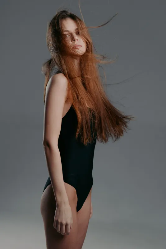Young woman with long hair wearing black swimsuit posing against gray background in studio environment.