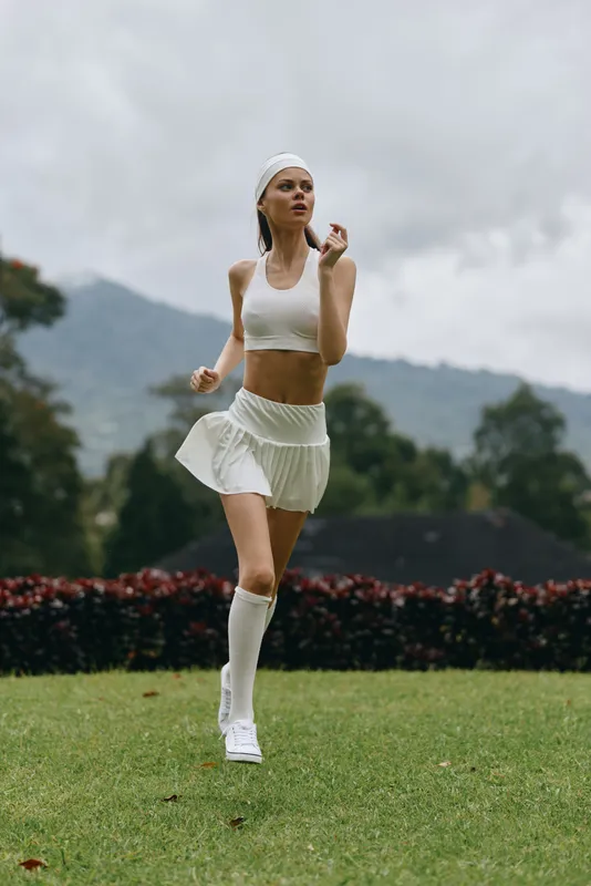 Athletic young woman in white sportswear jogging outdoors, showcasing health and fitness against a lush green background, conveying a vibrant and energetic lifestyle