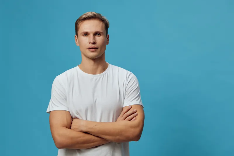 Young serious caucasian man in white t-shirt crossed arms look camera isolated on blue background digital nomad concept