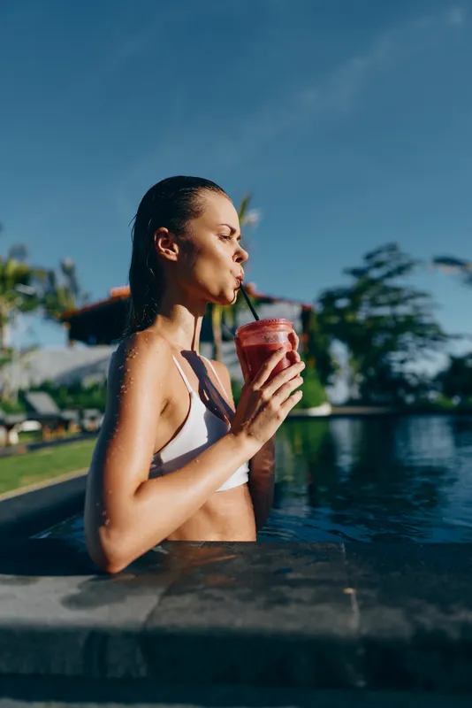 Smiling woman in a white bikini sipping a red drink at the pool edge. Bright outdoor setting with blue sky. Lifestyle moment emphasizing refreshment, relaxation, and wellness after a sunny day by the