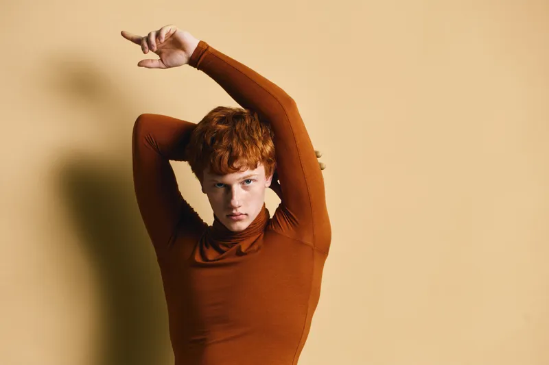 Portrait of young redhead woman posing with arms raised wearing brown turtleneck sweater on beige background with serious expression and direct gaze.