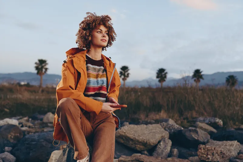 Woman lifestyle outdoors curly hair jacket standing sunset rocky nature, smiling with relaxed pose, orange jacket over striped sweater, hand gesture, soft evening breeze on rocky landscape