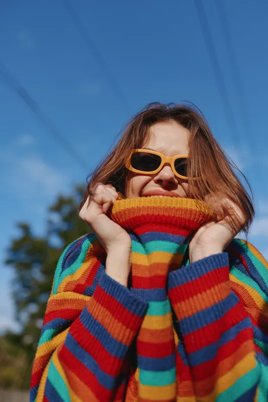Woman in a colorful striped sweater and sunglasses, portrait outdoors showing smiling expression and cozy casual style as she pulls the knit pullover up around her face.