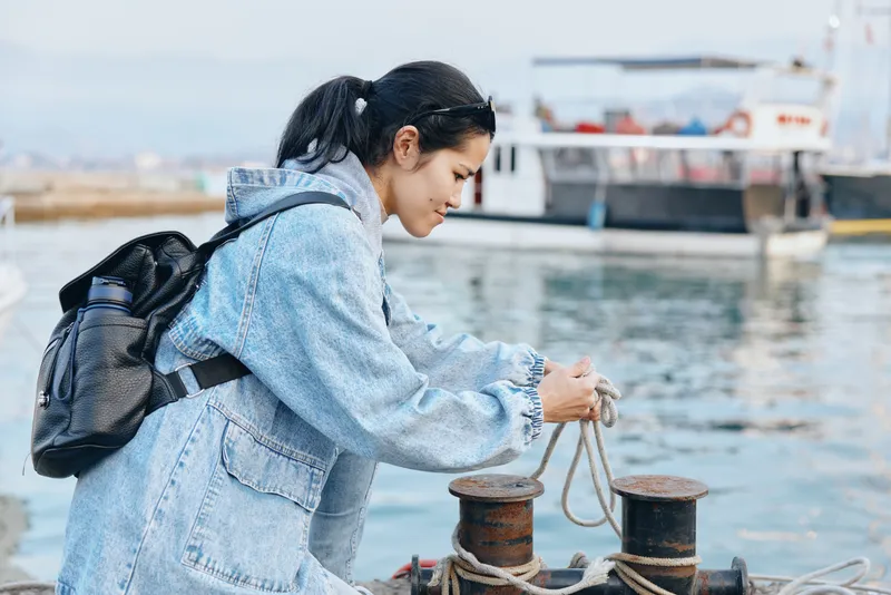 Woman in denim jacket tying rope at a seaside dock, beside mooring bollards and a calm harbor, with a backpack, suggesting travel, exploration and maritime work by the water.