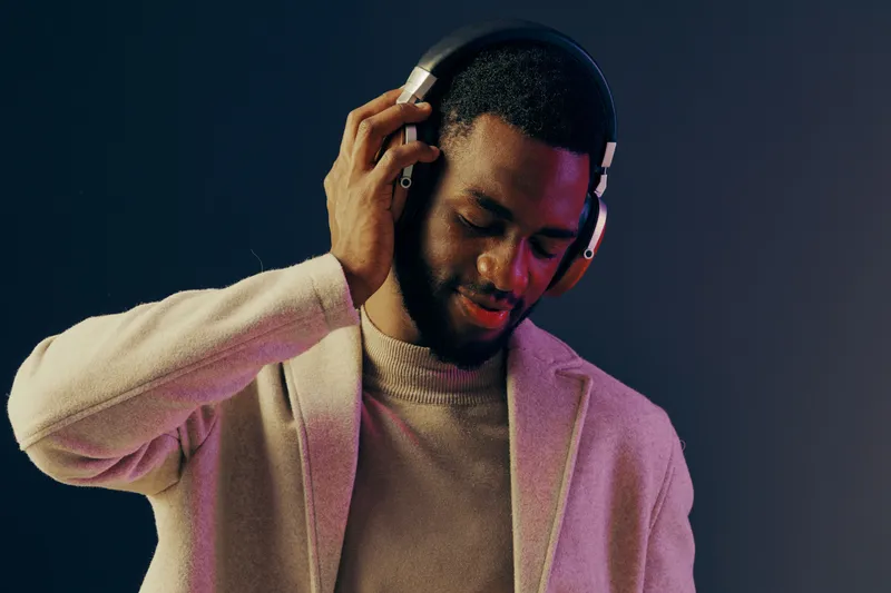 Young Black man enjoying music with headphones, wearing a stylish beige jacket and a sweater, creating a relaxed atmosphere with vibrant colors