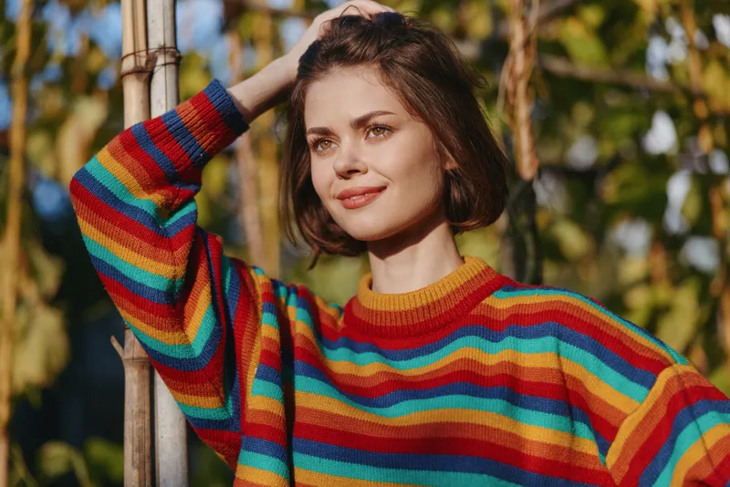 Woman smiling in a colorful striped sweater, short hair portrait leaning on bamboo pole outdoors in a sunlit garden. Casual outfit, relaxed mood for lifestyle and travel.