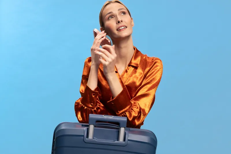 Young woman in orange silk blouse holds a suitcase and a smartphone, looking upwards with a hopeful expression. Travel concept, communication, planning, and anticipation in studio.