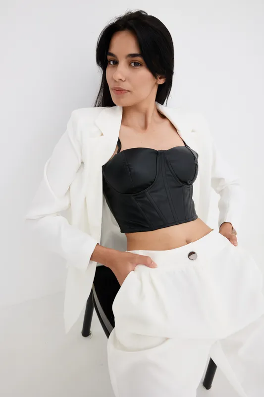 Stylish woman wearing black corset and white suit with matching pants, posing confidently against plain white background, showcasing modern fashion and elegance.