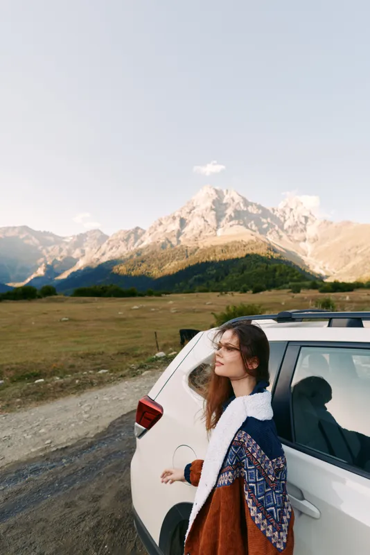 Woman car mountains road travel landscape: young woman leans on a white SUV beside a mountain road, cozy sweater and soft sunlight over valley and peaks during an outdoor road trip.