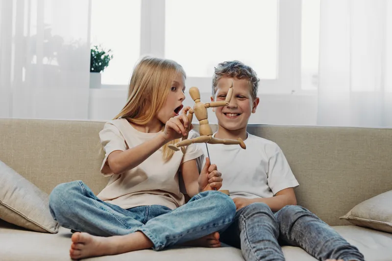 Two children sitting on a sofa play with a wooden mannequin, smiling and having fun in a bright living room with natural light and cozy atmosphere.