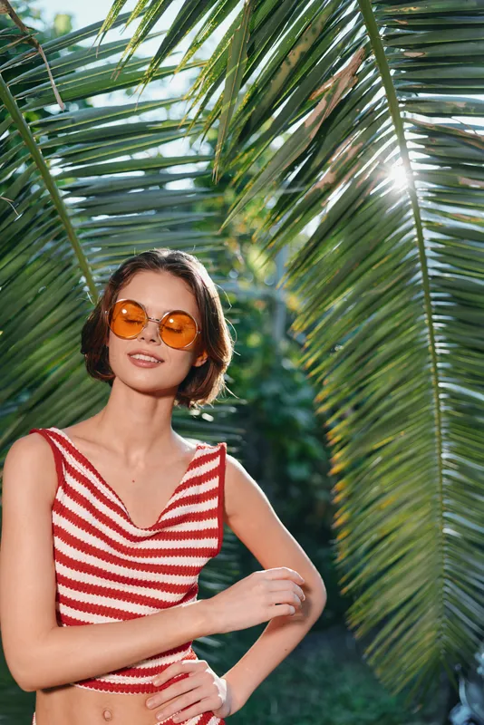 A cheerful young woman stands beneath palm fronds, wearing a red striped crop top and orange sunglasses, smiling as she enjoys bright outdoor light.
