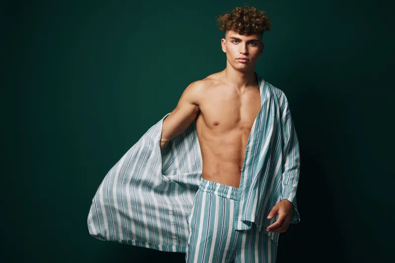 Young man with curly hair wearing striped pajamas posing against dark background, shirt open showing bare chest, casual and relaxed fashion style portrait.