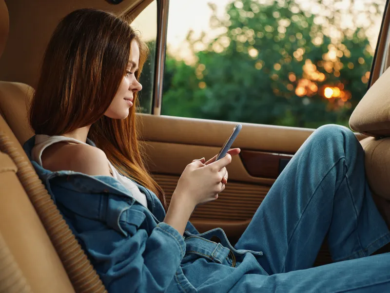 Young woman in denim outfit sitting in a car, using a smartphone, with a soft sunset background and greenery outside, evoking feelings of relaxation and connection.