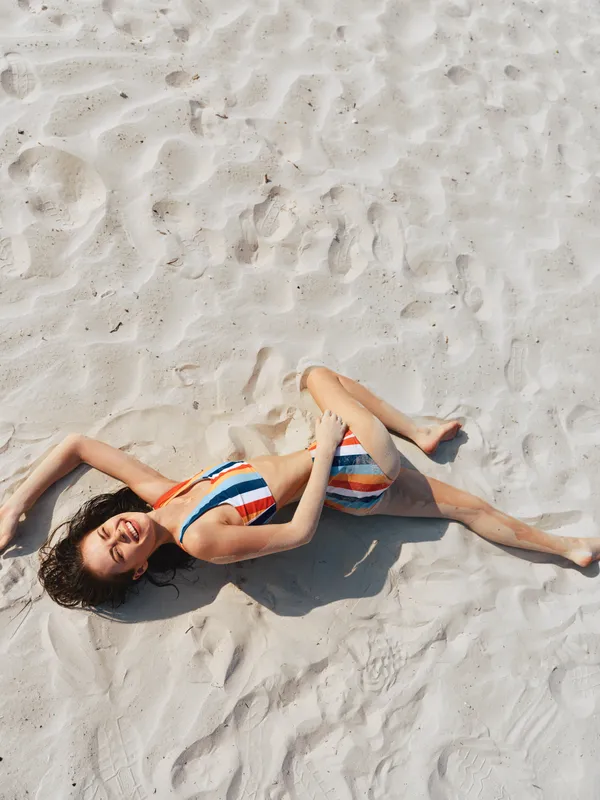 Sunlit scene of a woman in a striped bikini lying on soft sand, relaxed pose, highlighting vibrant swimwear and textured beach surface on a bright, carefree day.