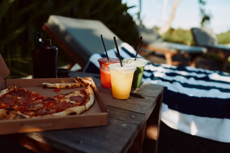 Casual outdoor meal with pizza on a wooden table and colorful drinks under sunny skies Relaxed lifestyle vibe with warm lighting capturing a social moment by the pool for friendly wellness and