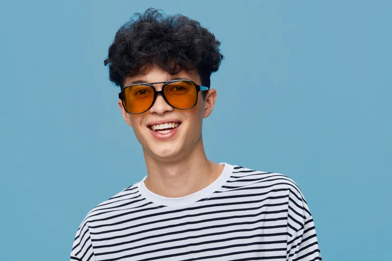 smiling man wearing yellow sunglasses and striped shirt on blue background, cheerful young person with curly hair expressing happiness and casual style