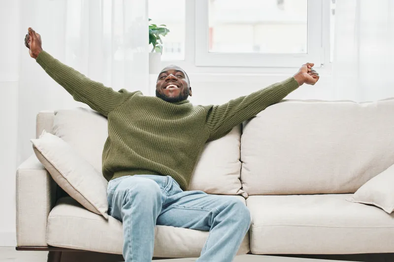 Happy Black man in a cozy green sweater relaxing on a couch, expressing joy and contentment in a bright and modern living room