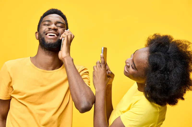 Joyful young African couple laughing, wearing yellow shirts, with vibrant yellow background, capturing moments on smartphones, expressing happiness and connection