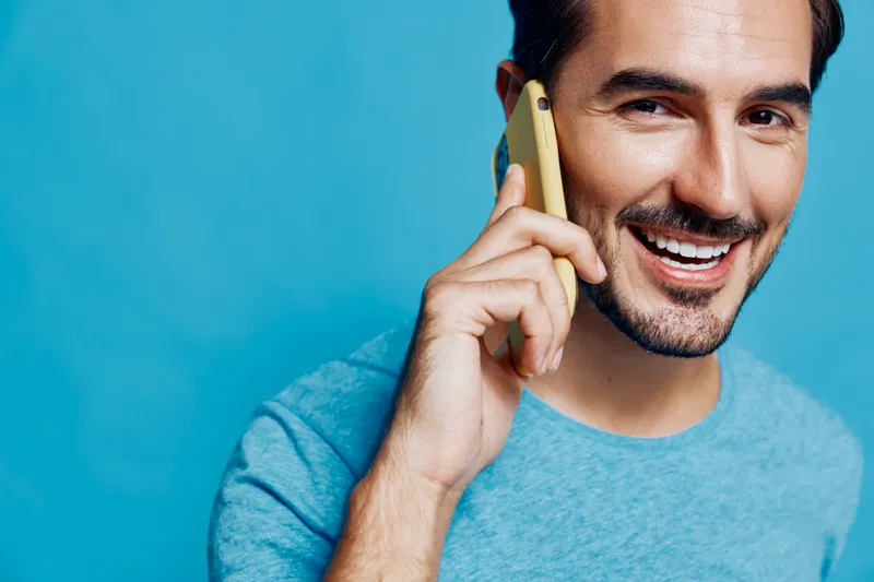 Smiling young man in a blue t-shirt chatting on his phone, showcasing confidence and approachability against a vibrant blue background. Lifestyle concept.